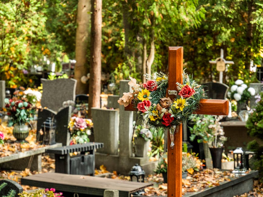 A wooden funeral cross decorated with a floral arrangement featuring red roses, yellow daisies, and white lilies, arranged with green foliage, placed among graves in a cemetery. The surrounding area s