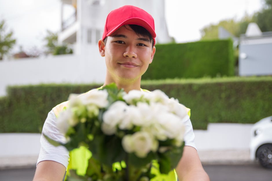 A delivery person wearing a red cap and a neon yellow vest holds out a bouquet of fresh white roses and green foliage towards the camera, smiling. The bouquet is wrapped in clear plastic with a green
