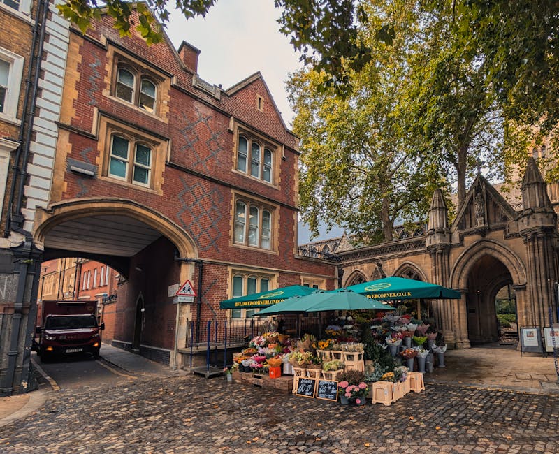 A vibrant outdoor flower market located on a cobblestone street beside a historic brick and stone building with arched passageways. The stall is sheltered under a green umbrella with the website 'flow