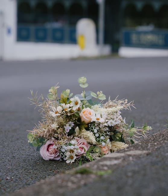 A delicate floral bouquet resting on the edge of a paved street, consisting of soft pink and white roses, small white daisies, and greenery including eucalyptus and dried, silvery foliage. The arrange