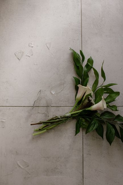 A small bouquet of fresh white calla lilies with smooth, trumpet-shaped petals and dark green, elongated leaves, arranged with some stems tied together with a natural twine. The bouquet rests on a lig