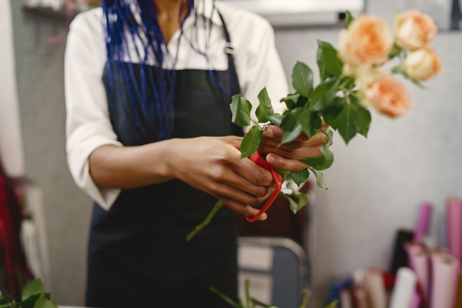 A florist in a white shirt and dark apron holds a bouquet of pale peach and cream roses with green foliage, secured with a red ribbon. The arrangement is in a glass vase or wrap, positioned at chest l