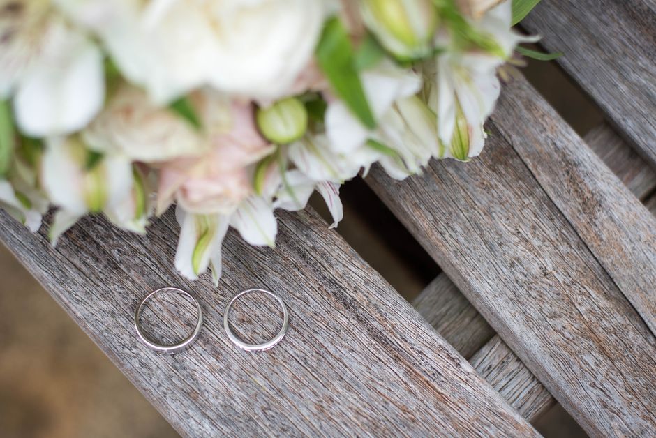 A pair of simple silver wedding bands placed on a weathered wooden surface beside a bouquet of fresh, pastel-colored flowers that include white and light pink petals with hints of green foliage. The b