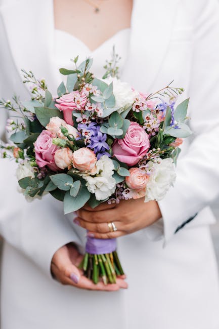 A person dressed in a white jacket holds a beautifully arranged bouquet of fresh flowers, featuring pastel pink roses, white lisianthus, lavender hyacinths, and sprigs of eucalyptus with muted green l
