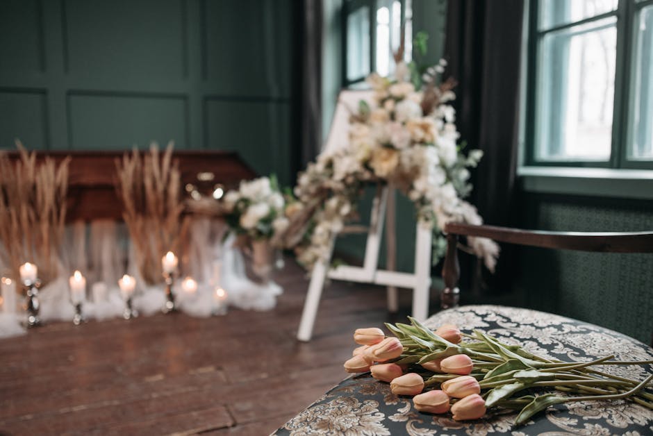 A floral arrangement featuring soft pink tulips with green stems rests on an ornate patterned chair in a softly lit room, with a large white and cream bouquet on a stand and a floral display with crea