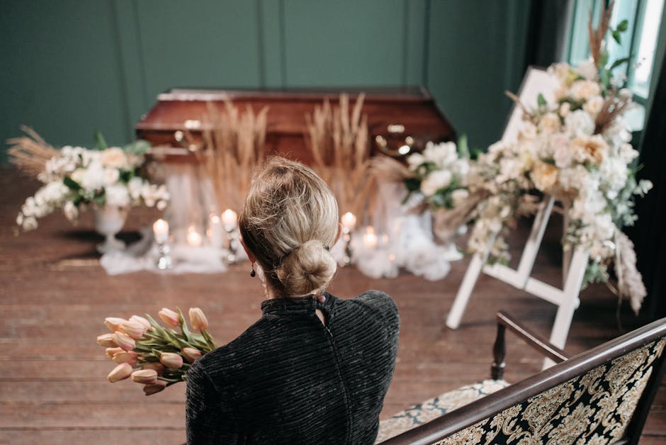 A woman with blonde hair styled in a low bun, wearing a dark, textured jacket, is seated with her back to the camera, holding a bouquet of pink tulips. She faces a floral arrangement set up for a fune