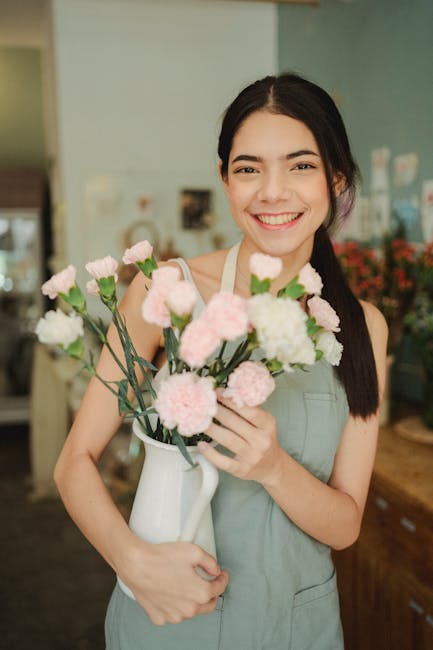 A young woman with dark hair pulled back, smiling warmly while holding a white ceramic vase filled with a bouquet of fresh carnations in shades of white and soft pink. She is wearing a light grey apro