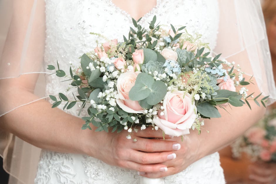 A bridal bouquet featuring soft pink roses, white baby’s breath, and eucalyptus leaves, arranged in a rounded style and wrapped with delicate white ribbon. The bouquet is being held by a bride dress