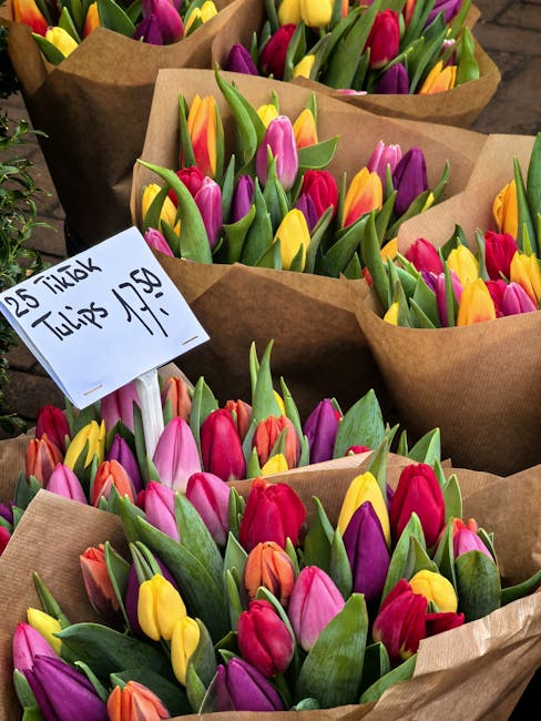A collection of freshly picked tulips arranged in brown paper wrapping, featuring vibrant colors including yellow, pink, purple, red, and orange. The tulips have smooth, slightly glossy petals and stu
