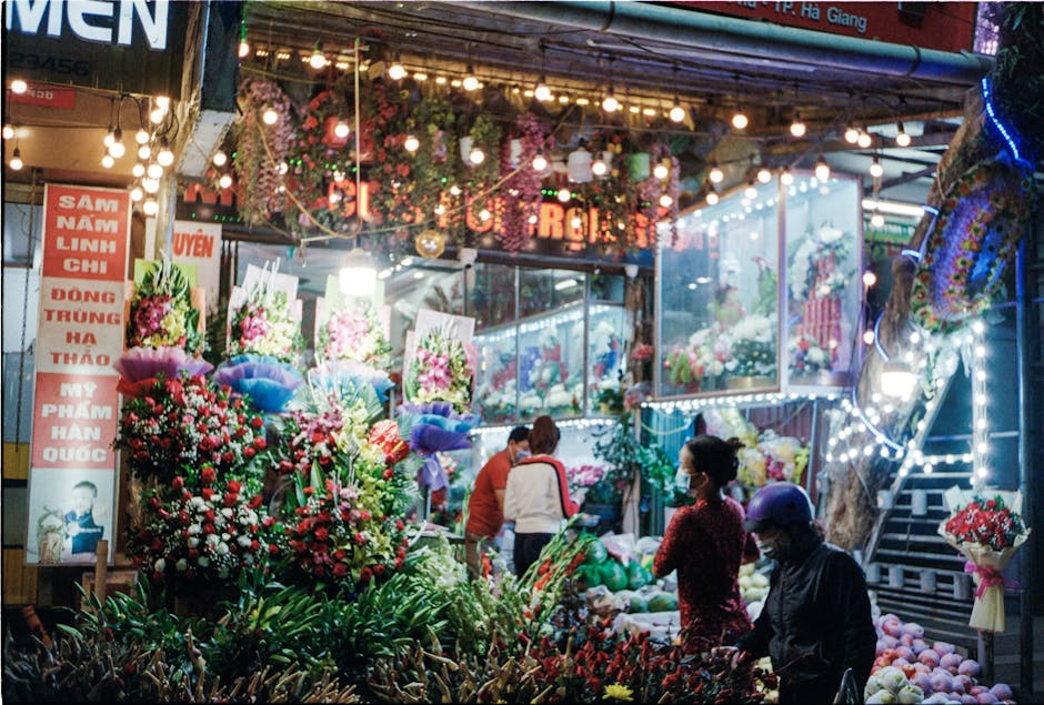 A vibrant flower stall on Mitcham High Street illuminated by decorative string lights, showcasing a variety of floral arrangements. The display includes bouquets of red, pink, and white flowers, with 