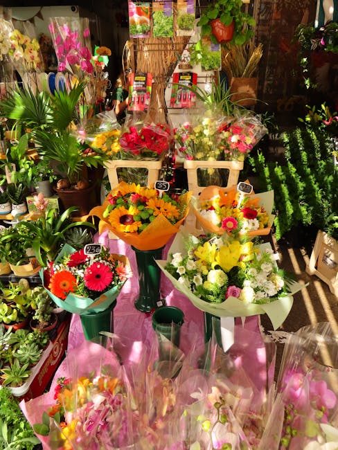 A vibrant display of fresh floral bouquets arranged on a pink cloth-covered table inside a flower shop. The bouquets feature a variety of flowers including bright pink gerbera daisies, yellow sunflowe